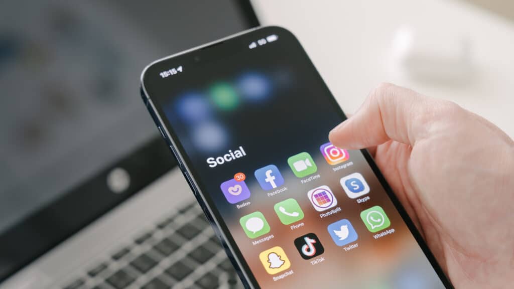 A man's hand holds a Iphone 13 Pro max with various social media apps, against the background of a laptop.